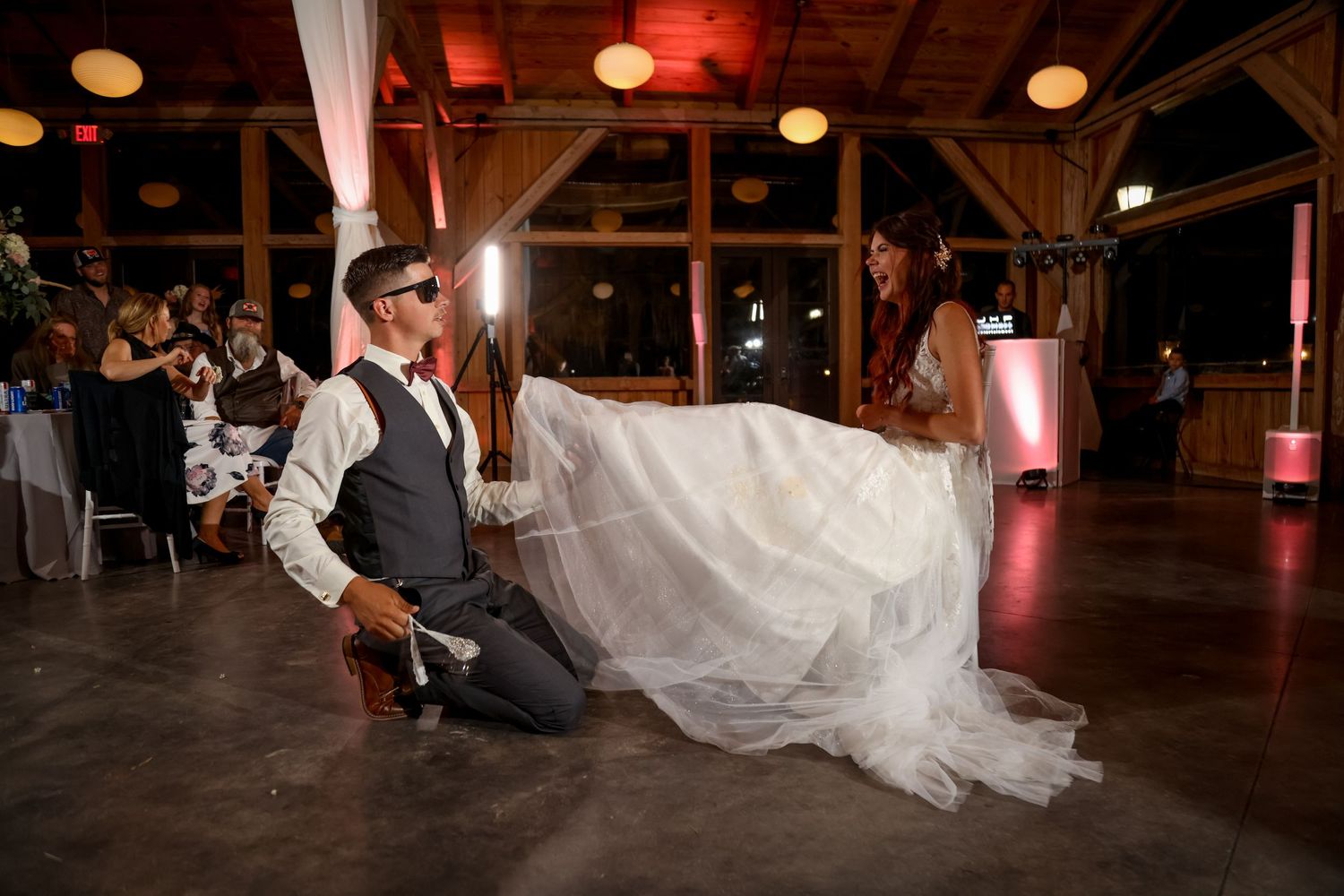 removing of the garter moment at a wedding reception where the groom wears sunglasses as bride laughs