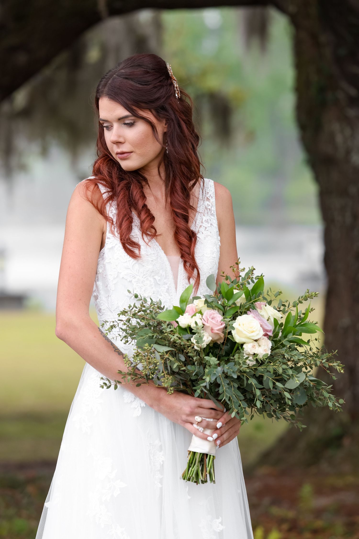 A bride in a white lace dress holds a romantic bouquet with blush roses and greenery in an outdoor garden setting.
