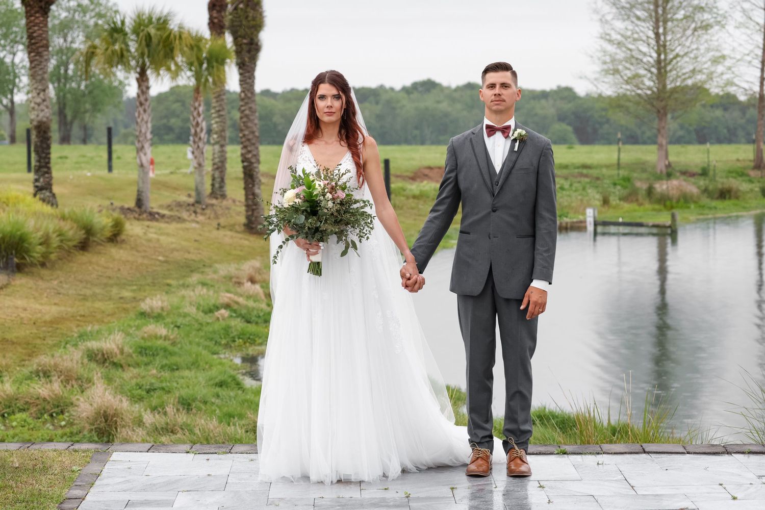 A couple holds hands on their wedding day next to a serene pond surrounded by palm trees and greenery.