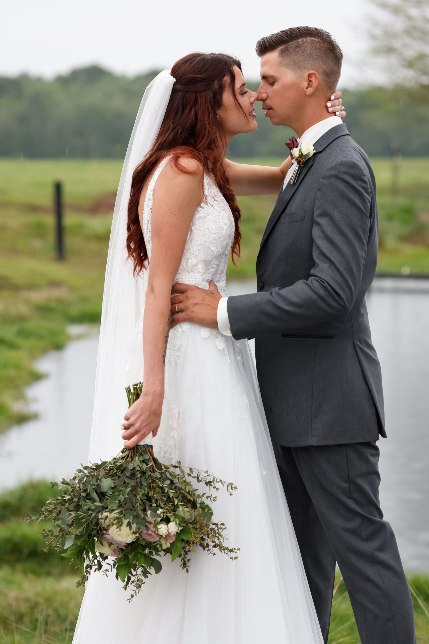 A bride in a white lace dress holds a green and floral bouquet while sharing an intimate moment with groom in grey suit outdoors.