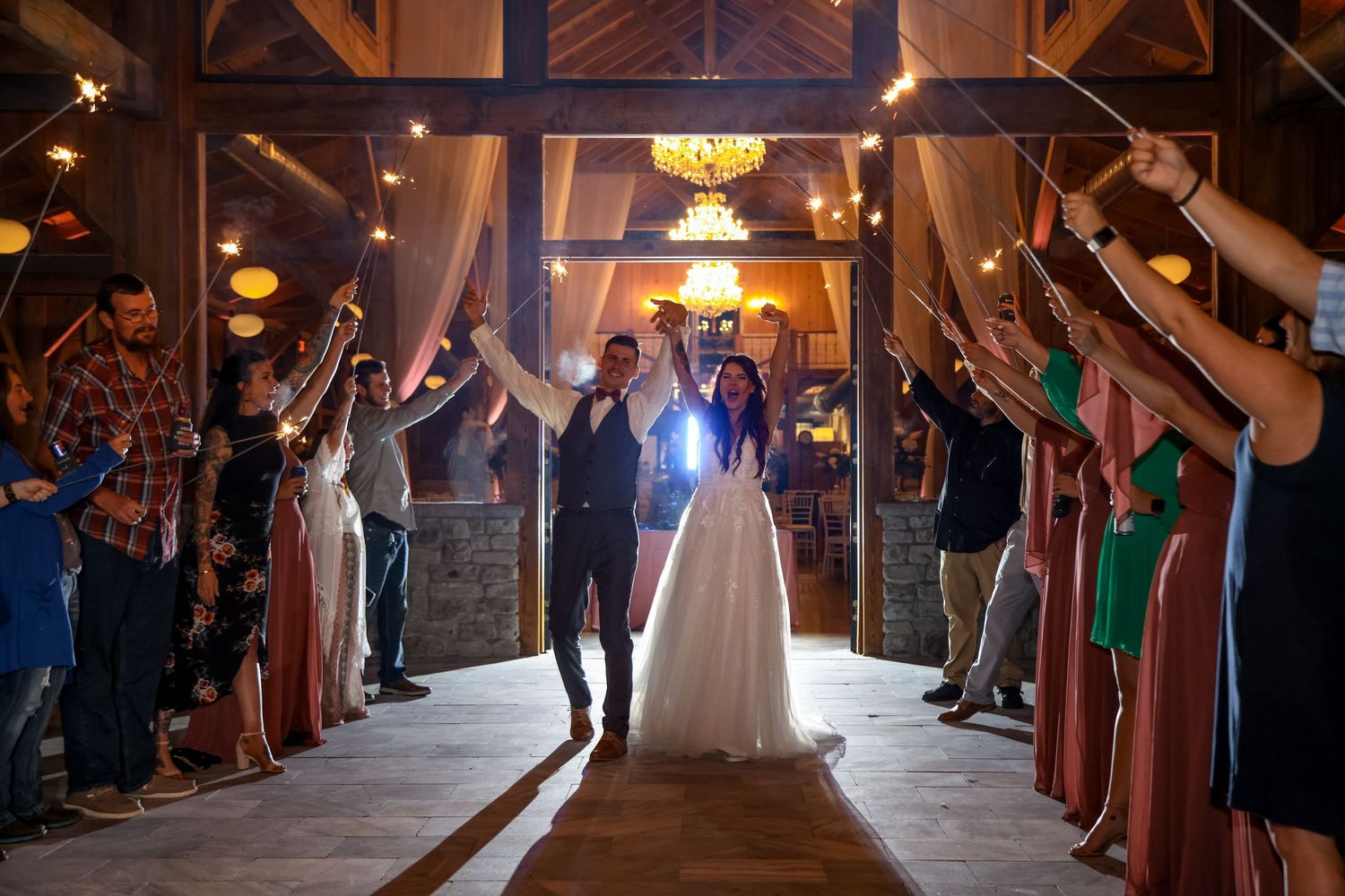 A newlywed couple exits their wedding reception through a tunnel of guests holding sparklers in a rustic wooden venue.