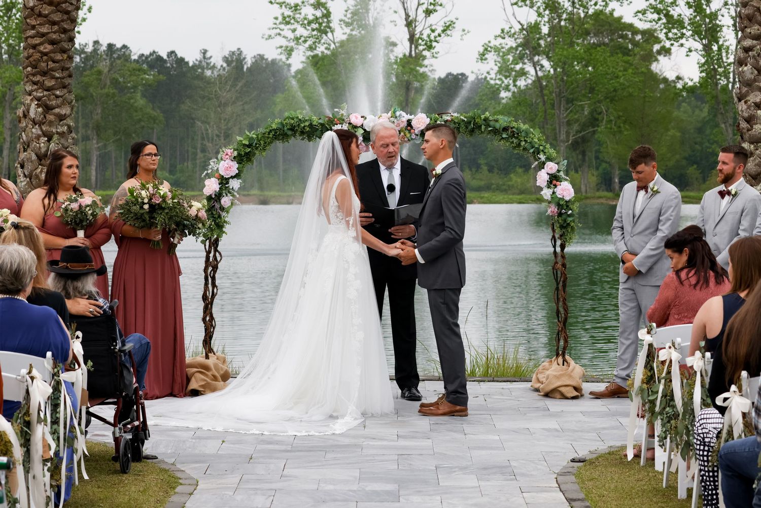 A picturesque lakeside wedding ceremony takes place on a stone patio with a fountain spraying in the background.