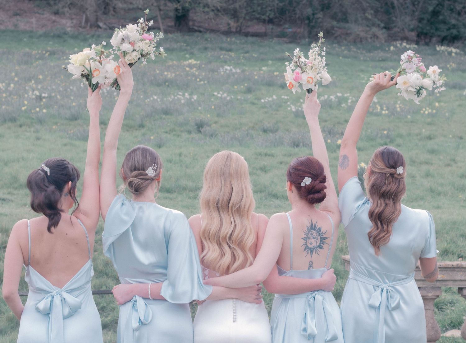 Bridesmaids in light colored dresses pose together outdoors with raised arms in celebration.
