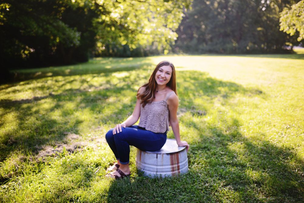A person in casual attire sits on a white bucket in a sunlit grassy field during golden hour.