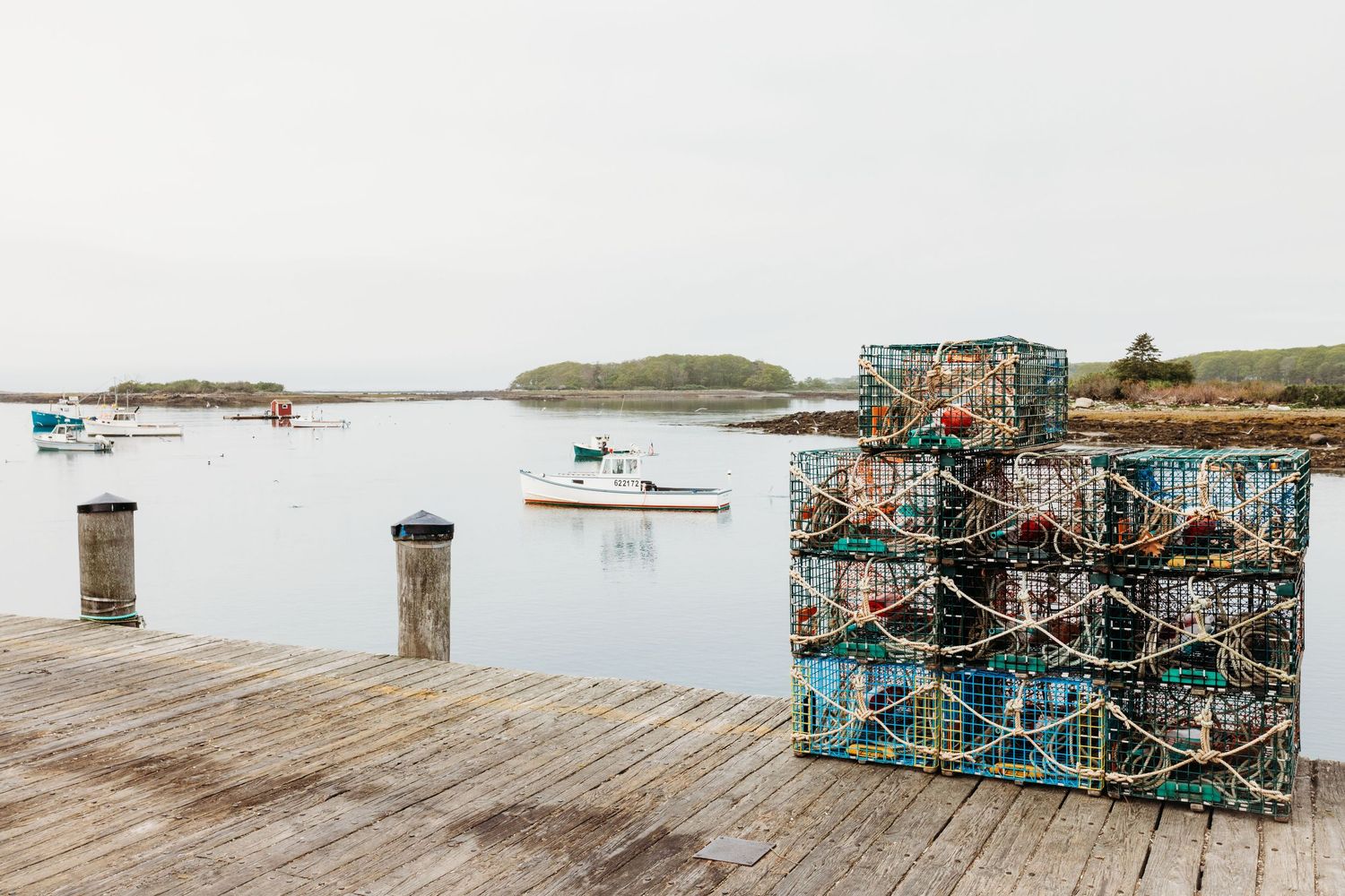 Wooden dock with stacked lobster traps and fishing boats in calm harbor waters.