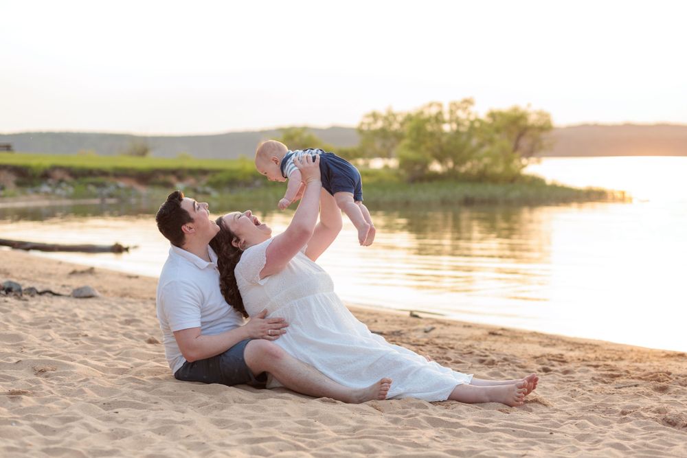 A couple sits on a sandy beach at sunset playing with a baby against a serene waterfront backdrop at Jordan Lake in Raleigh NC