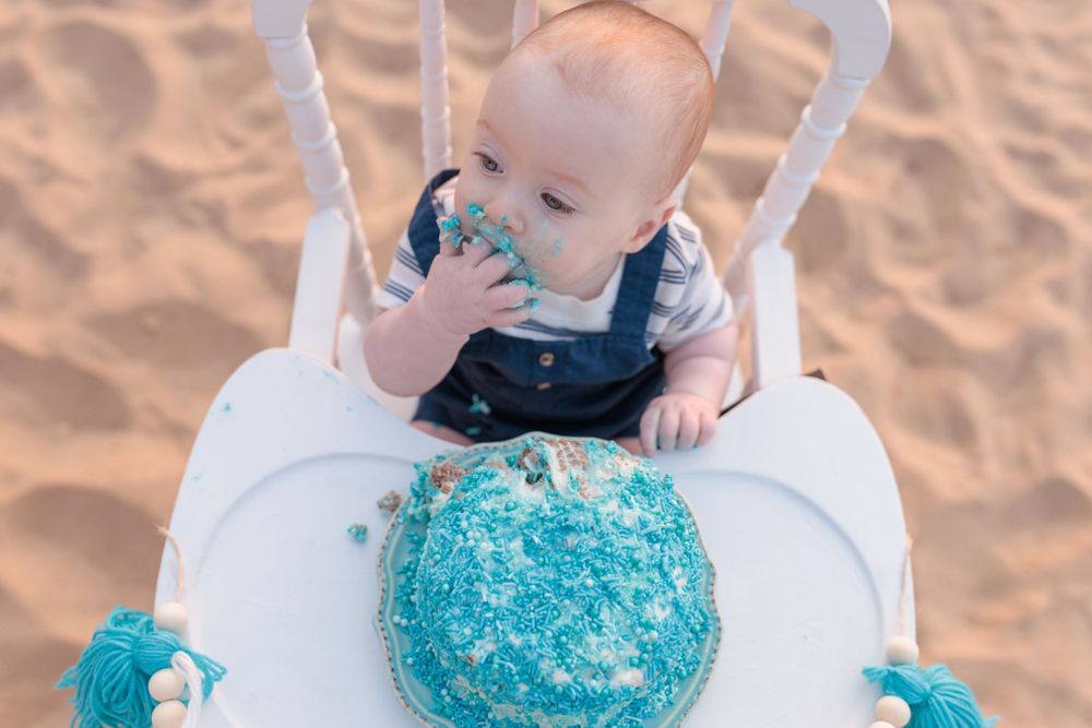 Baby sits in high chair with blue smash cake during first birthday celebration on sandy beach.