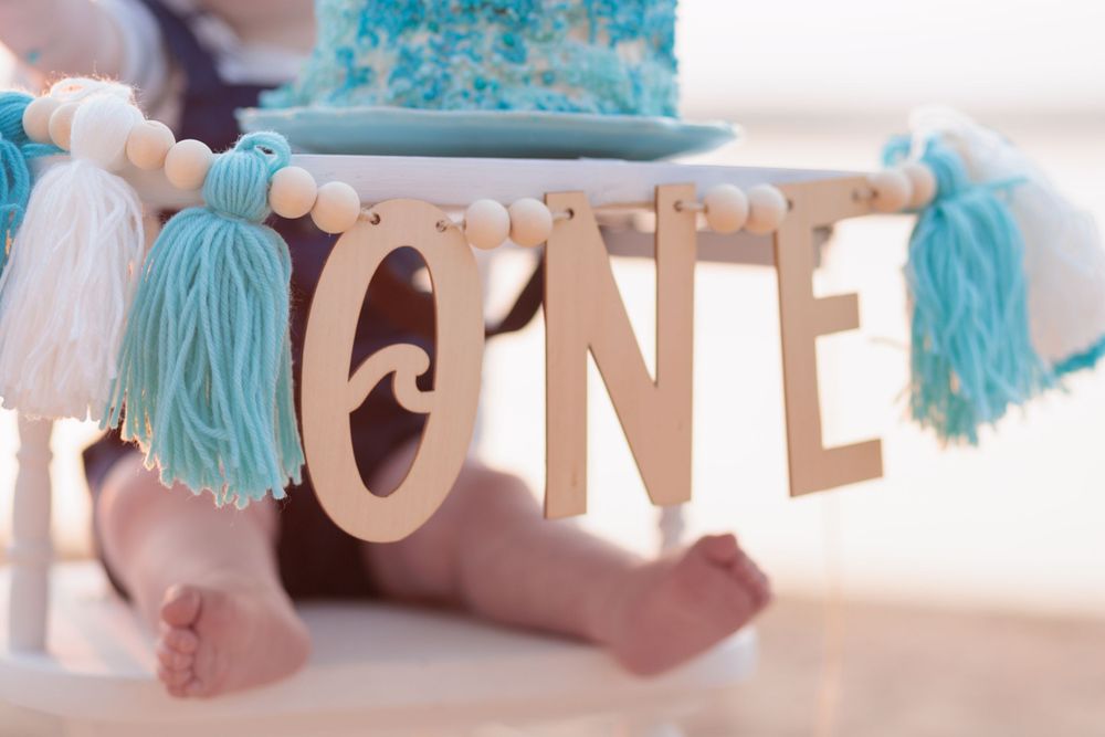 A wooden 'ONE' banner decorated with light blue tassels and white beads for first birthday party decor.