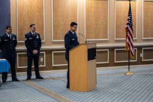 Service members participate in an indoor military ceremony with American flag displayed.
