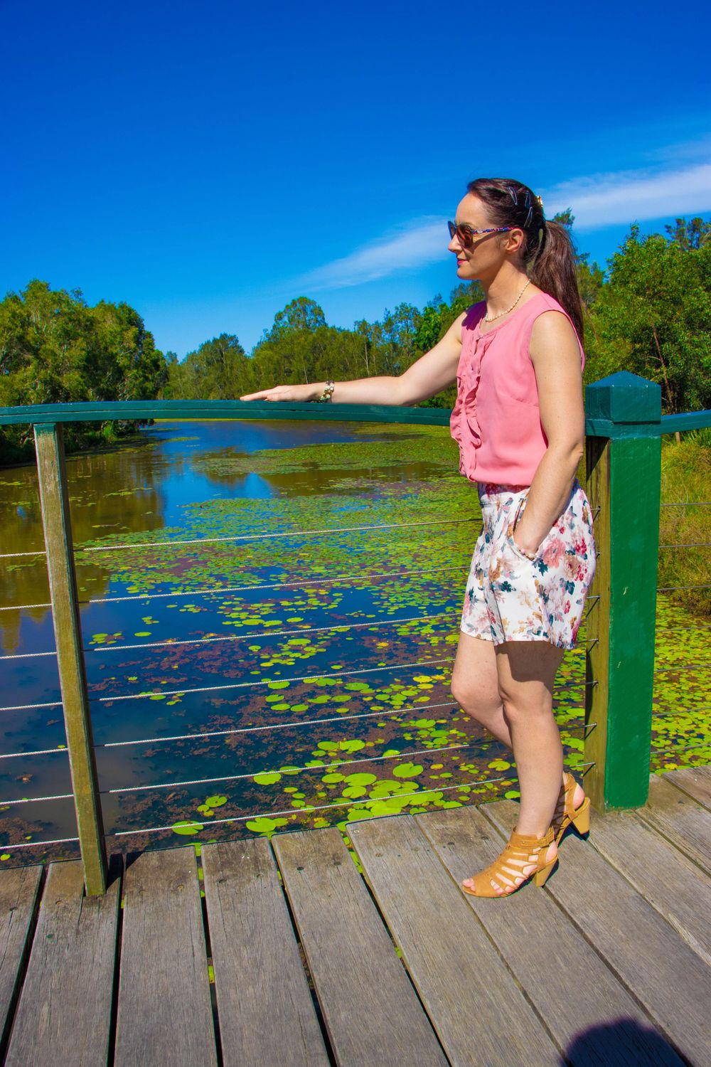 Person in pink top and floral shorts standing on wooden boardwalk overlooking pond with lily pads.