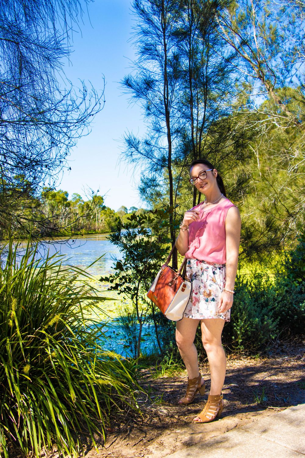 A sunny lakeside path with greenery and someone in a pink top and floral skirt carrying a bag.