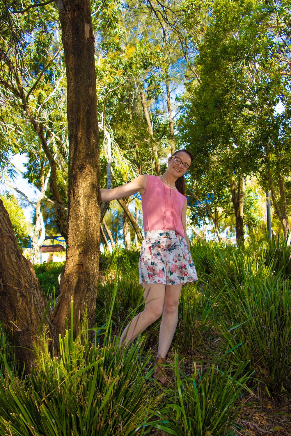 Person in pink top and floral shorts posing playfully among trees in sunlit park.