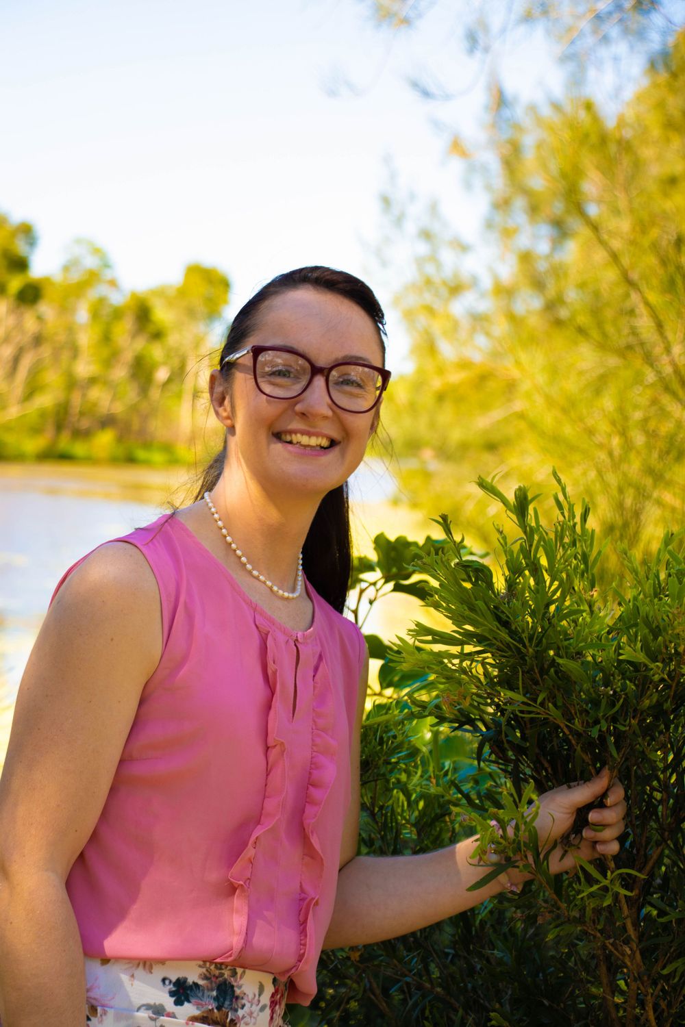 Person in pink sleeveless top holding green plant by waterside on sunny day.