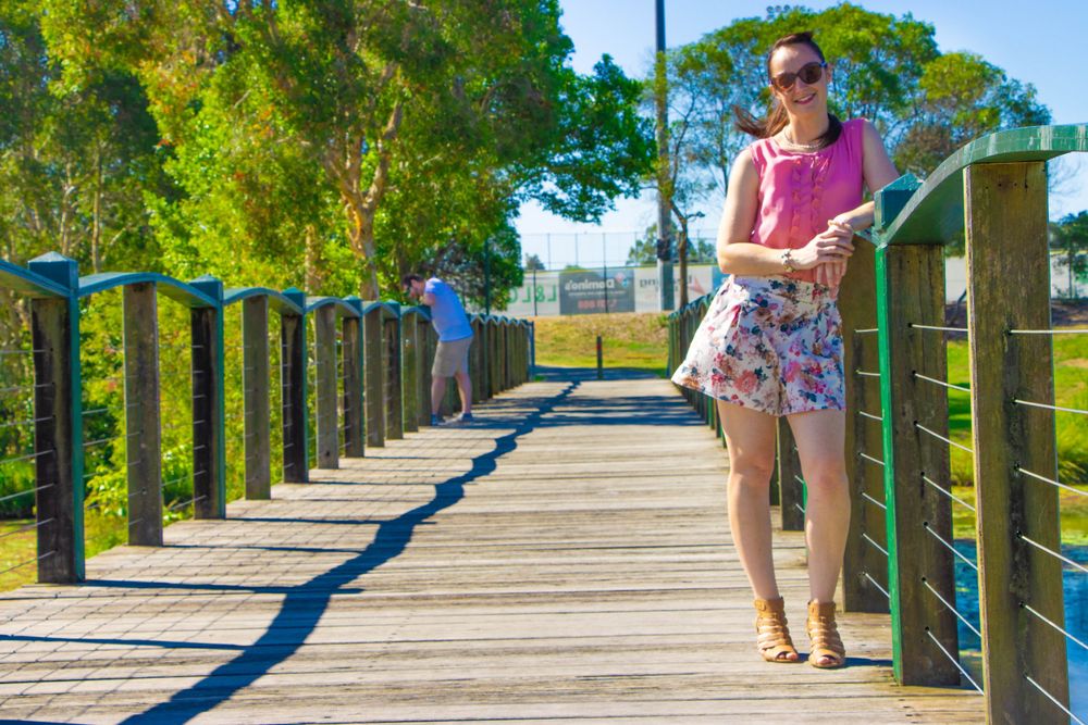 Person in pink shirt and floral shorts walking along a sunny wooden boardwalk over water.