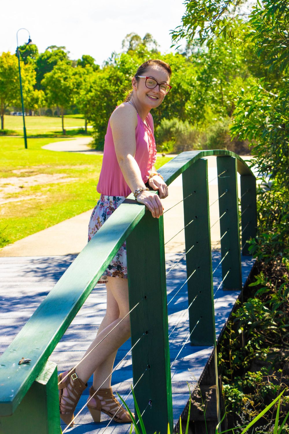Person in casual summer attire poses on green bridge railing with park scenery in background.