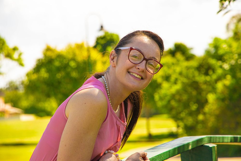 Person wearing glasses and pink top smiles while leaning on outdoor railing.