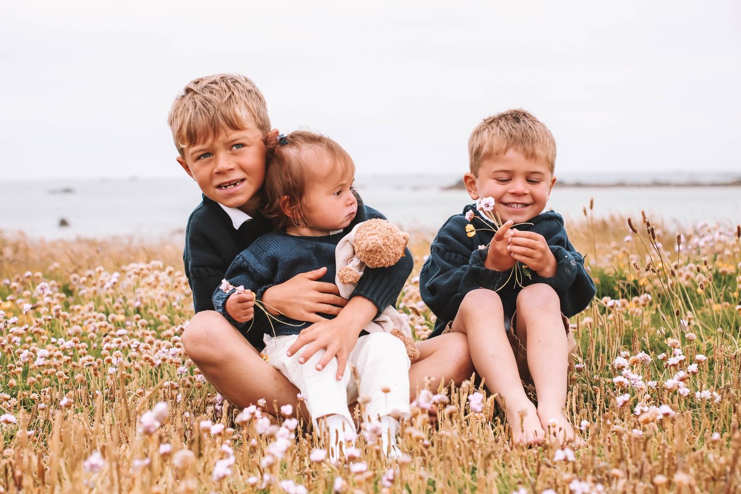 Three young children sitting together in wildflower field near coastline.