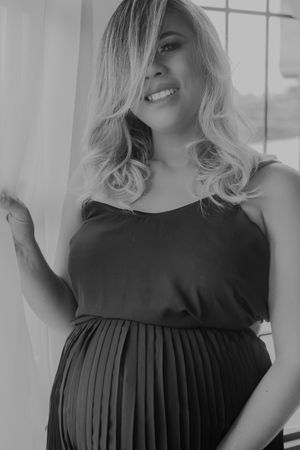 Black and white portrait of expectant mother in pleated dress standing by window with flowing curtains.