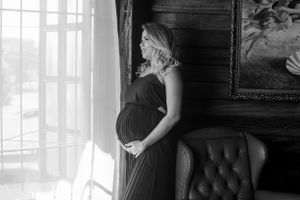 Black and white maternity photo with woman gazing through window by leather chair.