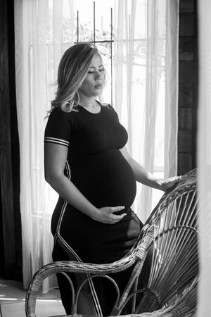 Black and white maternity photo showing woman in striped dress by window.