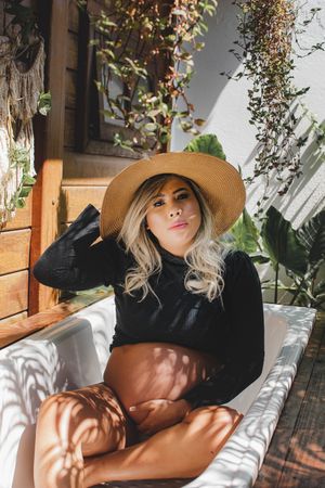 Person lounging on white cushions wearing black outfit and sun hat in garden setting.