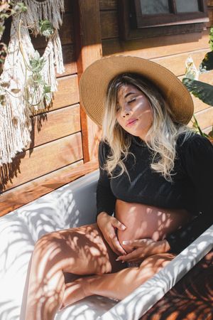 Woman in black attire and straw hat relaxing on white cushions with sunlight patterns.