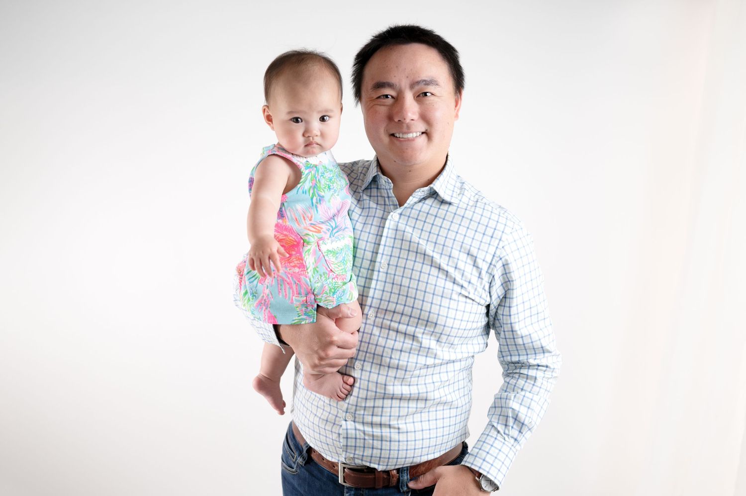 Adult in checkered shirt holds smiling baby wearing pink floral outfit against white background.