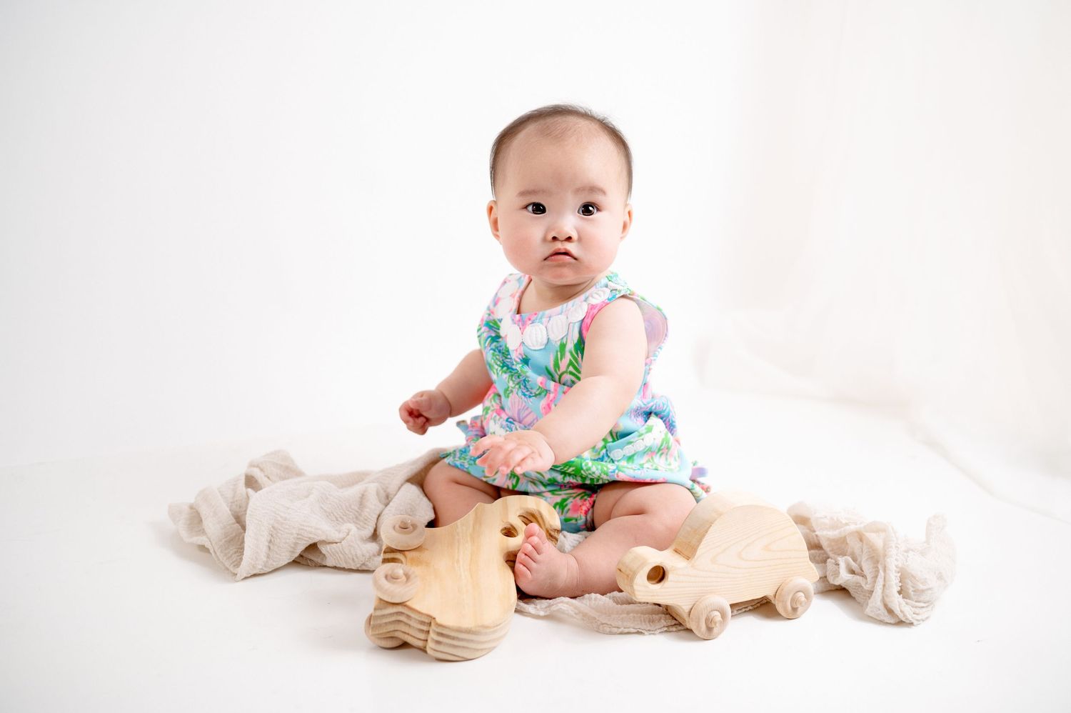 Baby in pastel outfit sits surrounded by plush toys on white background.