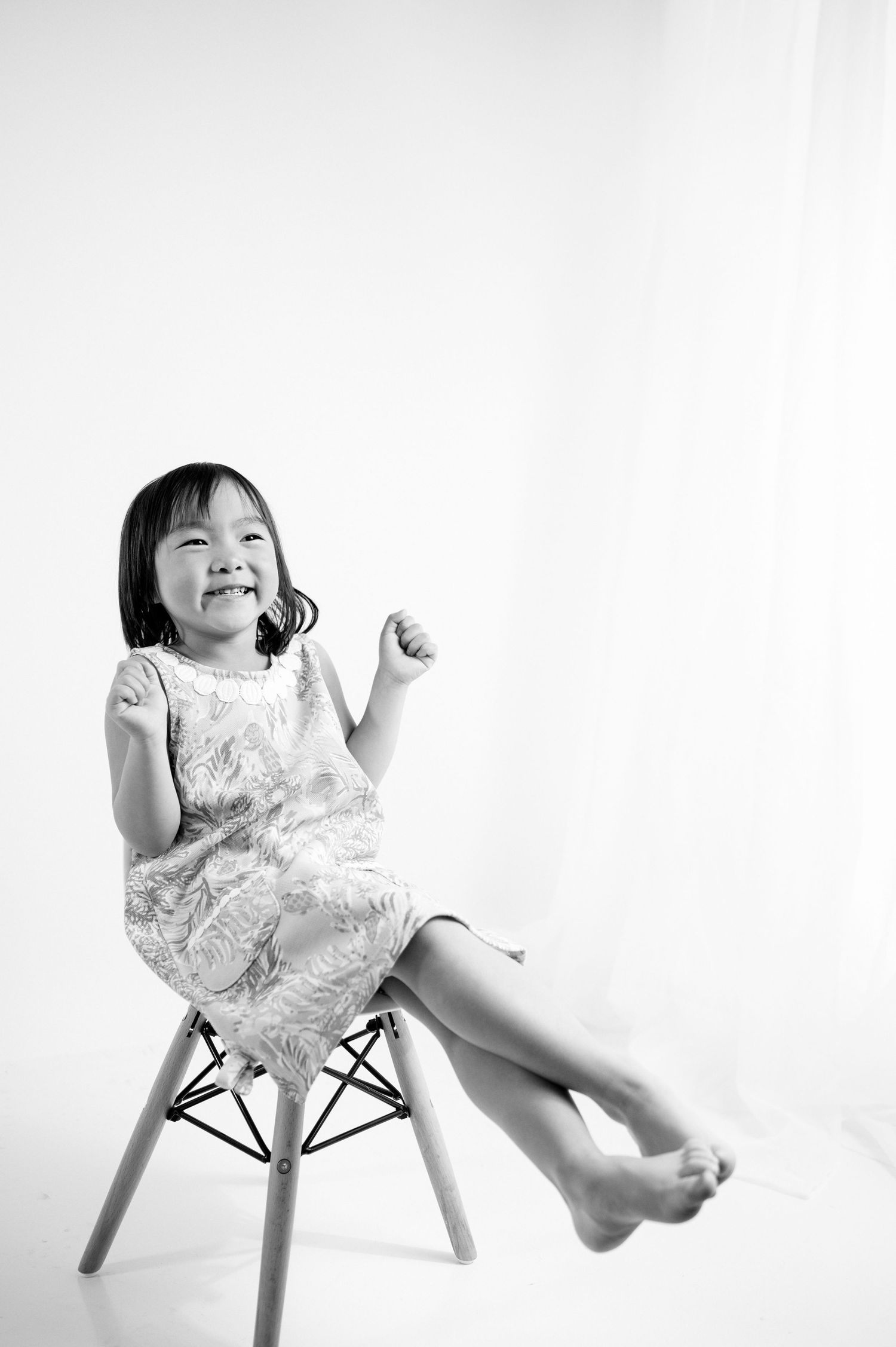 Happy child in white dress sits on modern chair with dangling feet against white background.