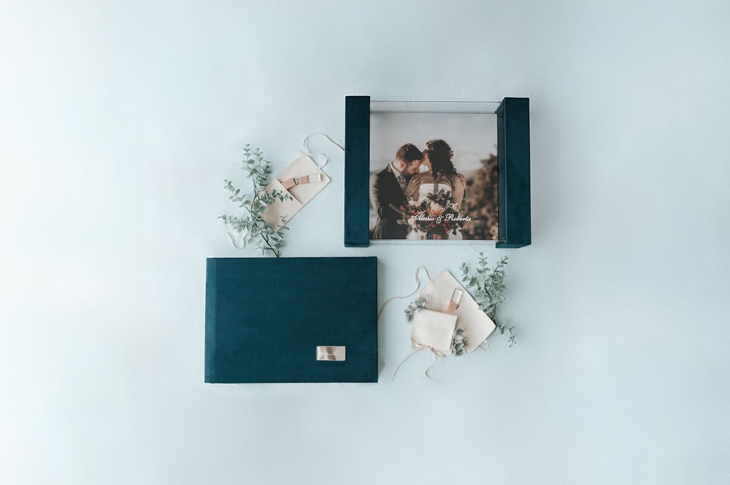 Teal velvet wedding album and open photo box with couple portrait surrounded by eucalyptus sprigs.