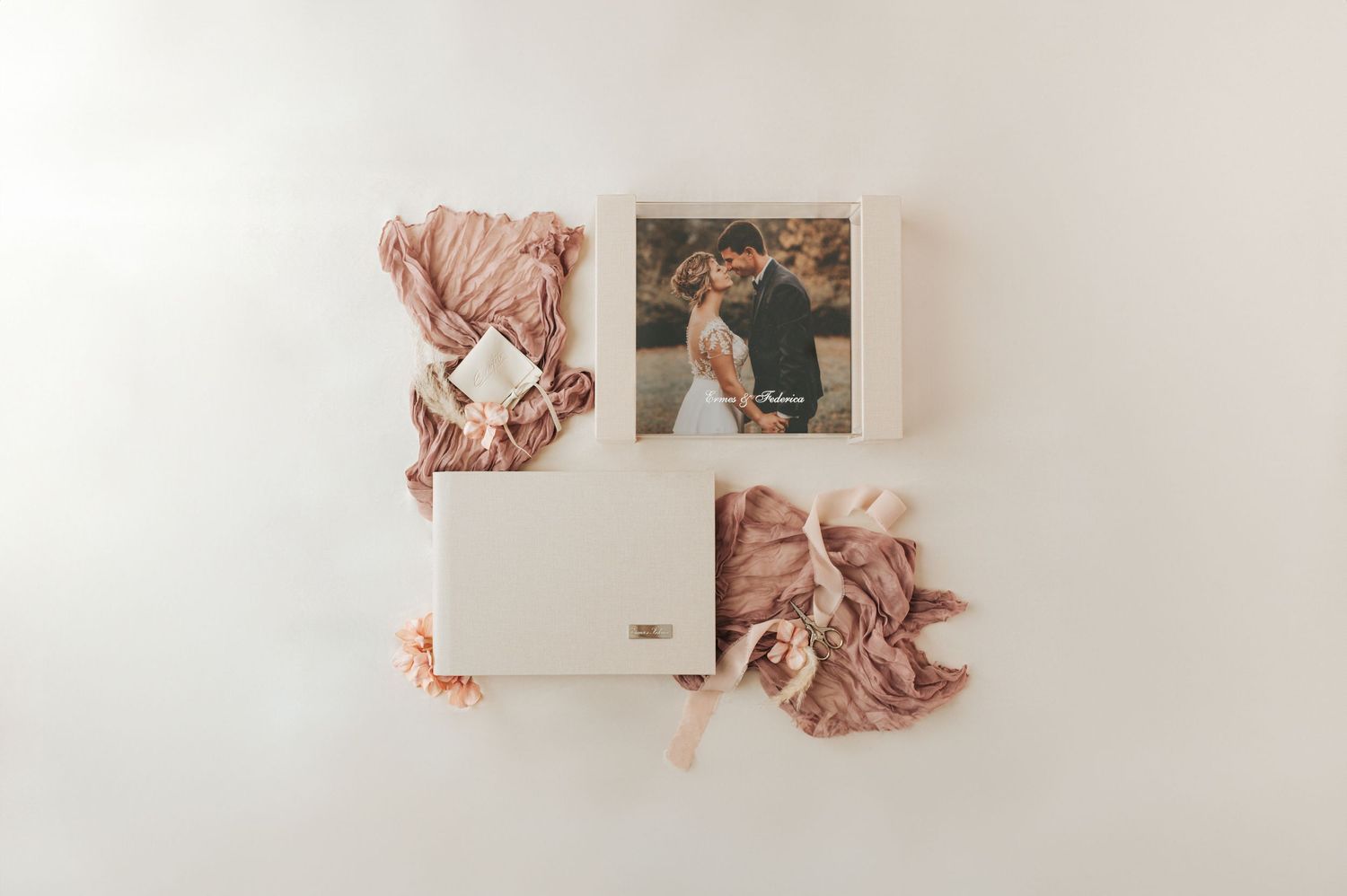 Flat lay of cream wedding photo album, printed couple portrait, pink silk ribbon, and blush floral accents on white.
