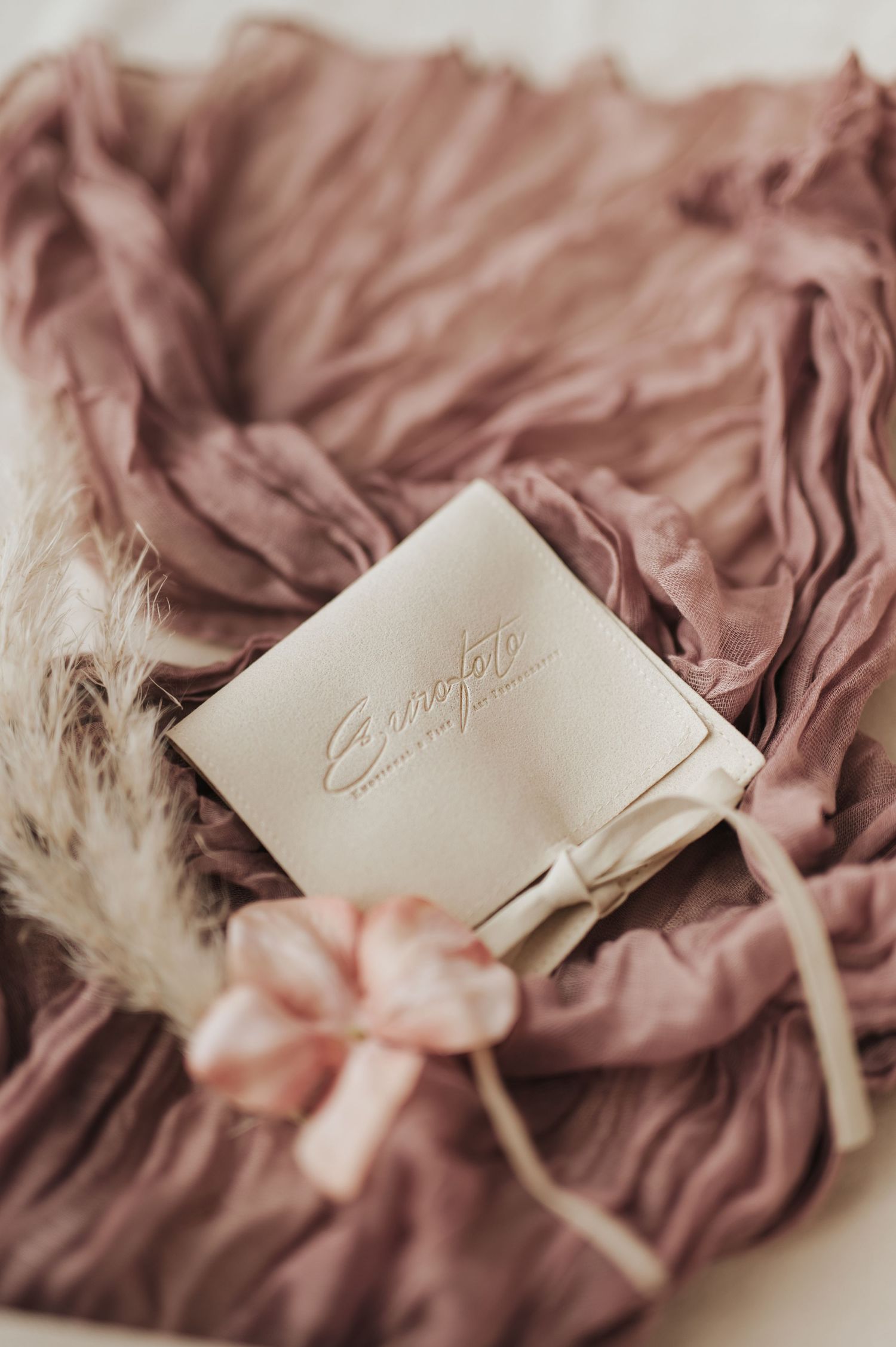 Cream velvet Enotete jewelry pouch with ribbon bow resting on dusty rose ruffled fabric with feathers and pink flowers.