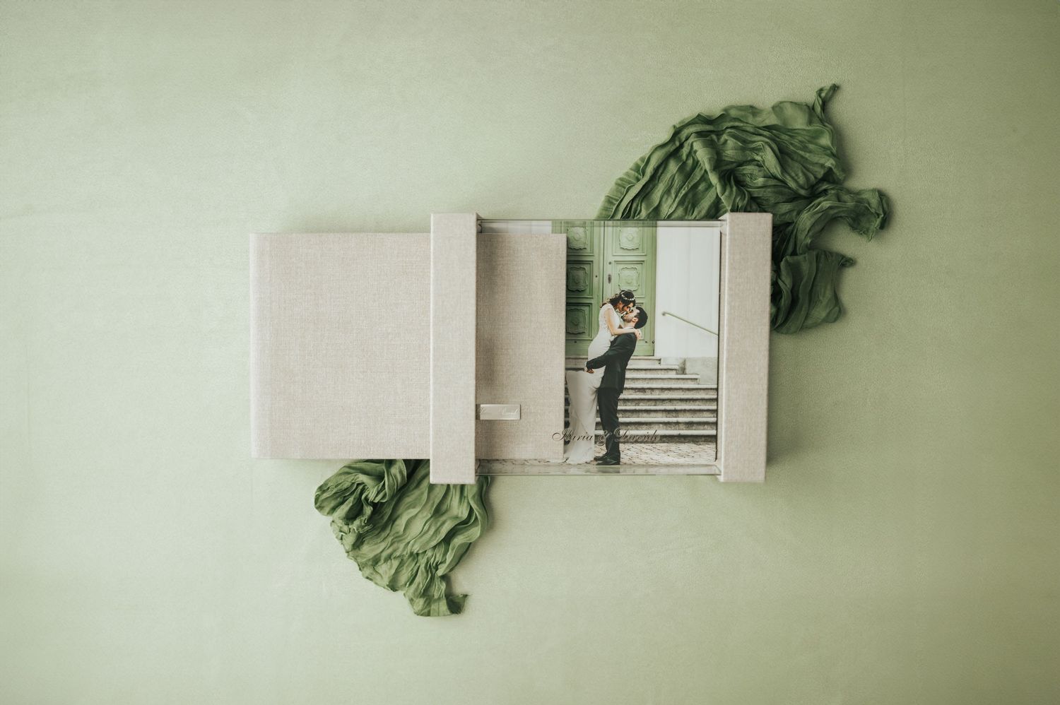 Open wedding photo album displaying a couple on stairs, with sage green fabric on green background.