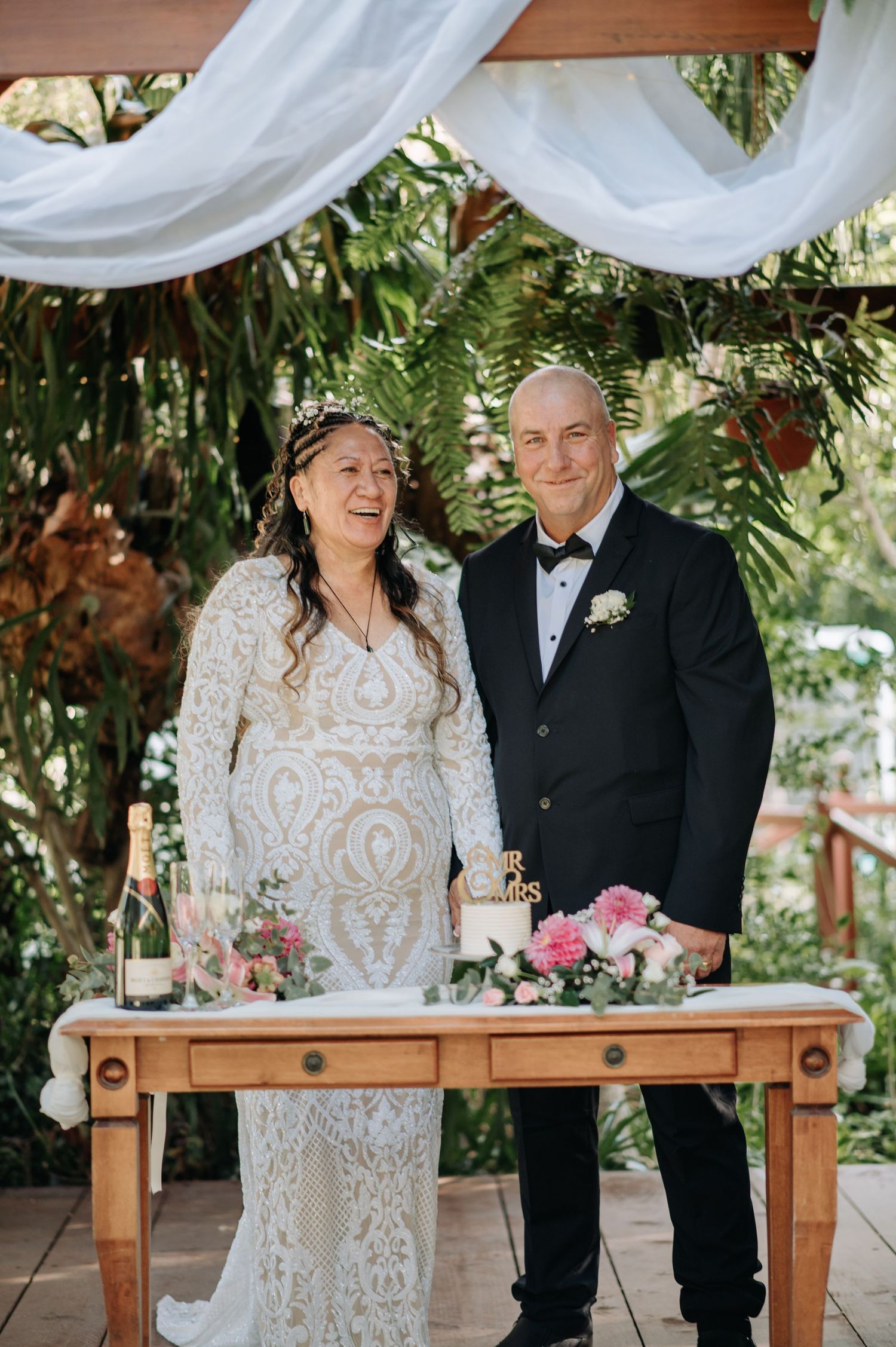 Couple shares joyful moment at rustic wooden ceremony table decorated with white fabric draping and greenery.