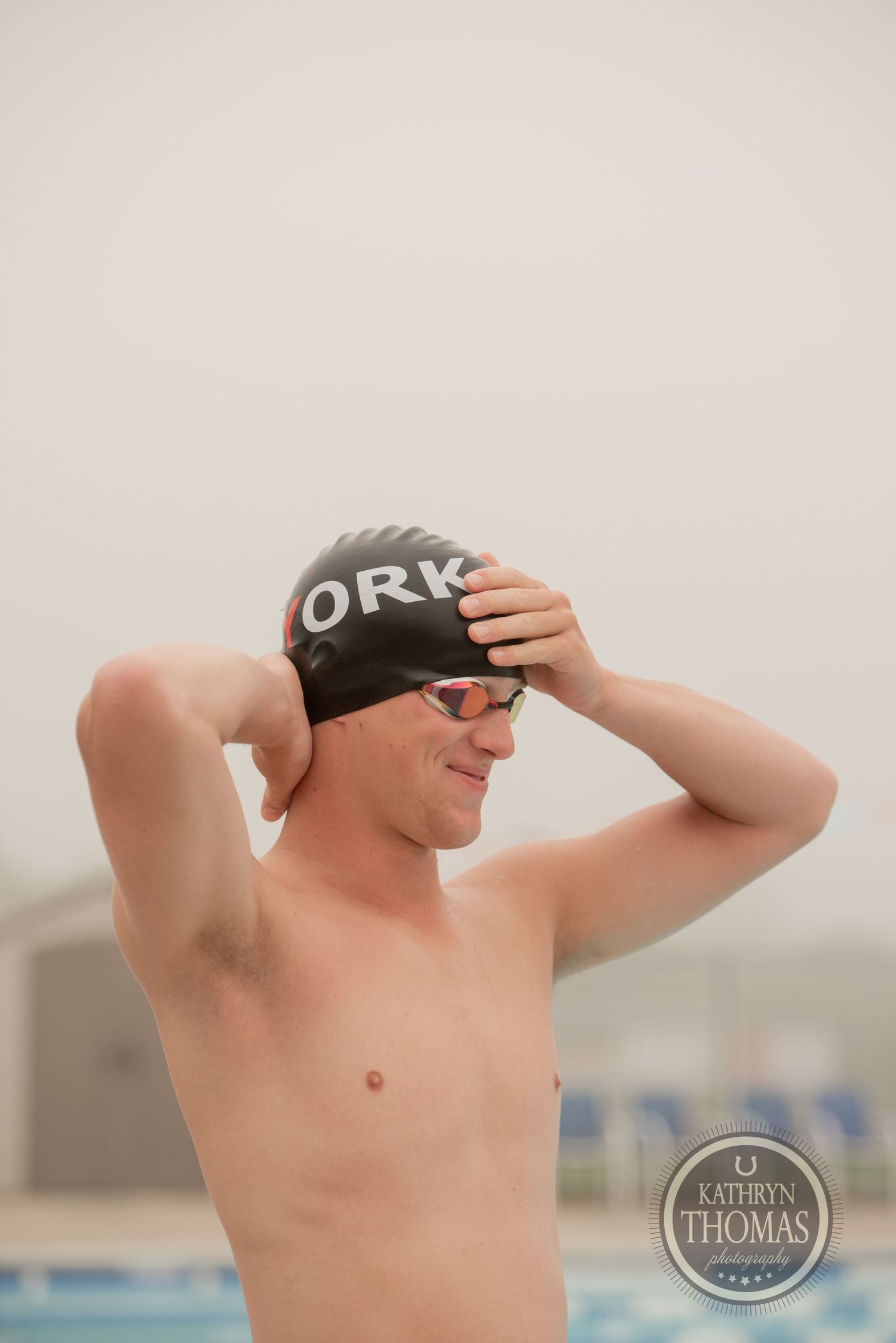 A shirtless swimmer adjusts their swim cap while standing poolside.