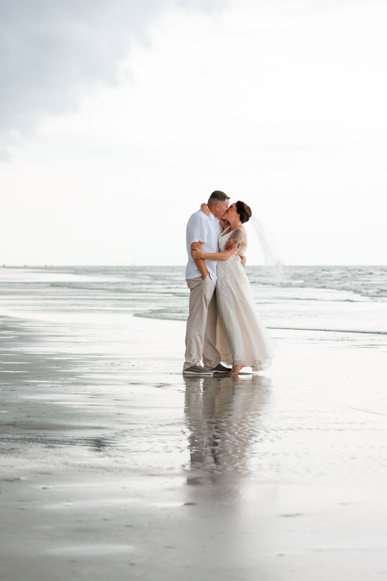 A romantic couple embraces on a misty beach shoreline while waves roll in on an overcast day.