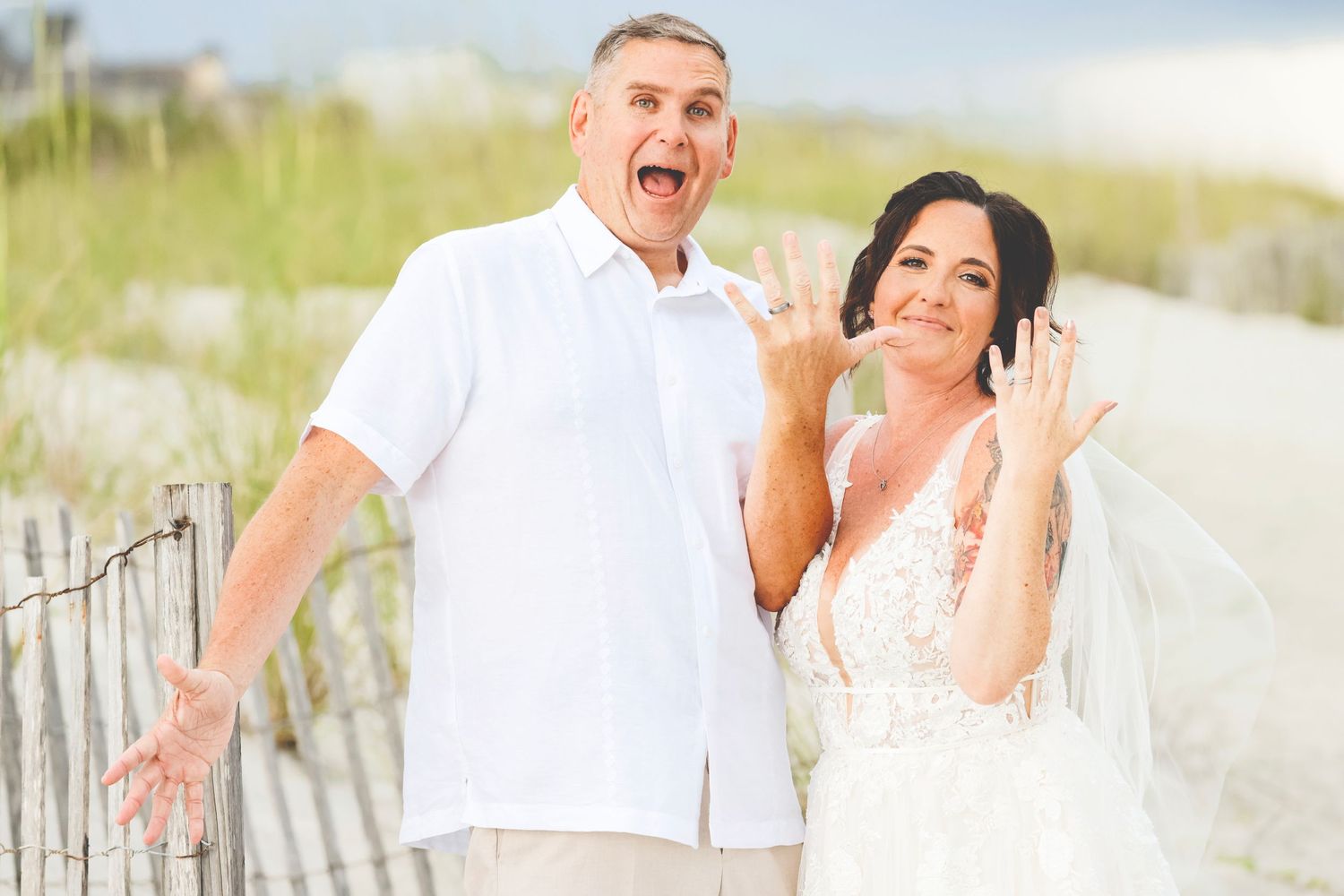 A couple pose playfully together on a sandy beach boardwalk, with the woman wearing a white lace dress and veil.