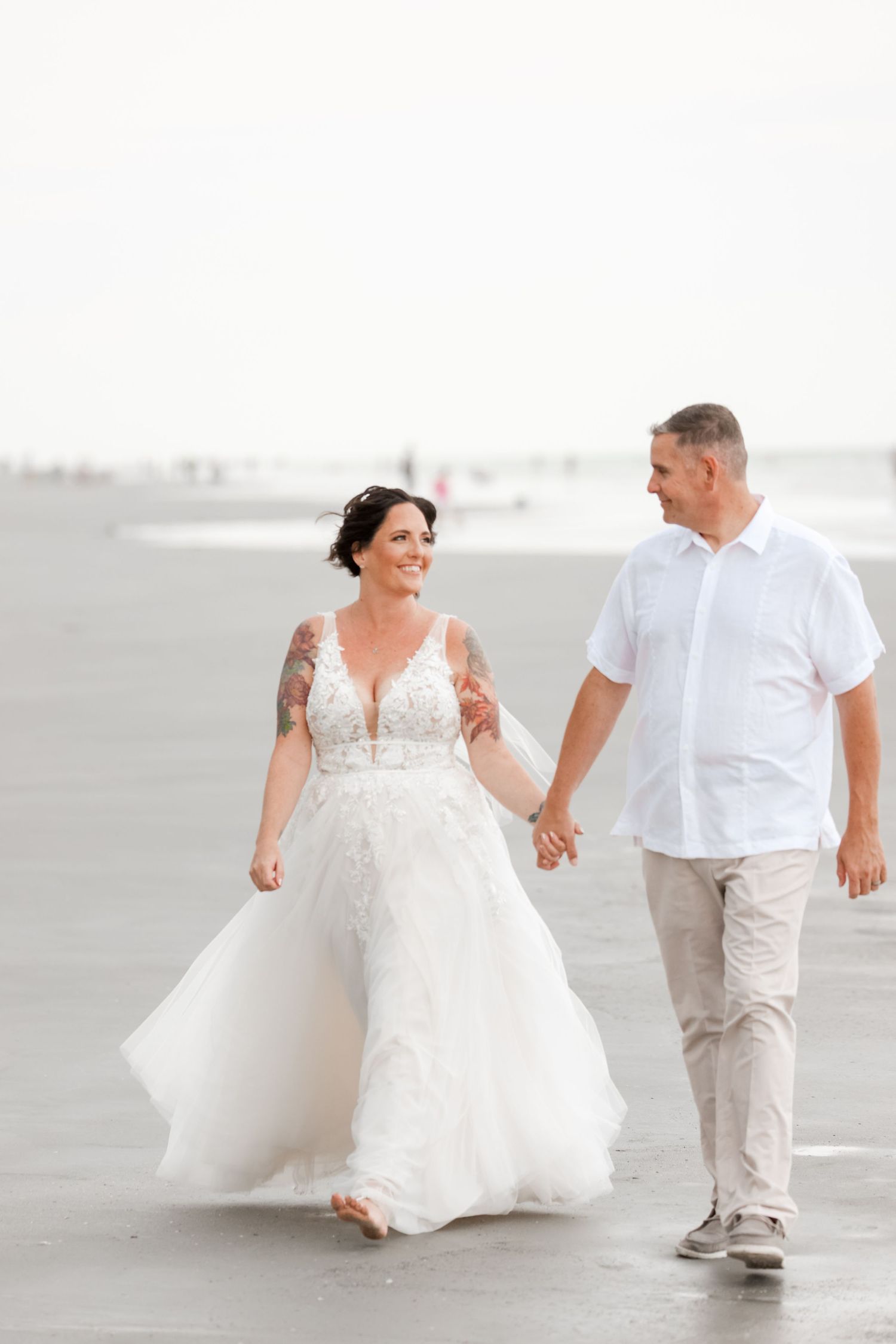 A couple walking hand in hand on a beach, with the bride wearing a flowing white gown and groom in casual white attire.