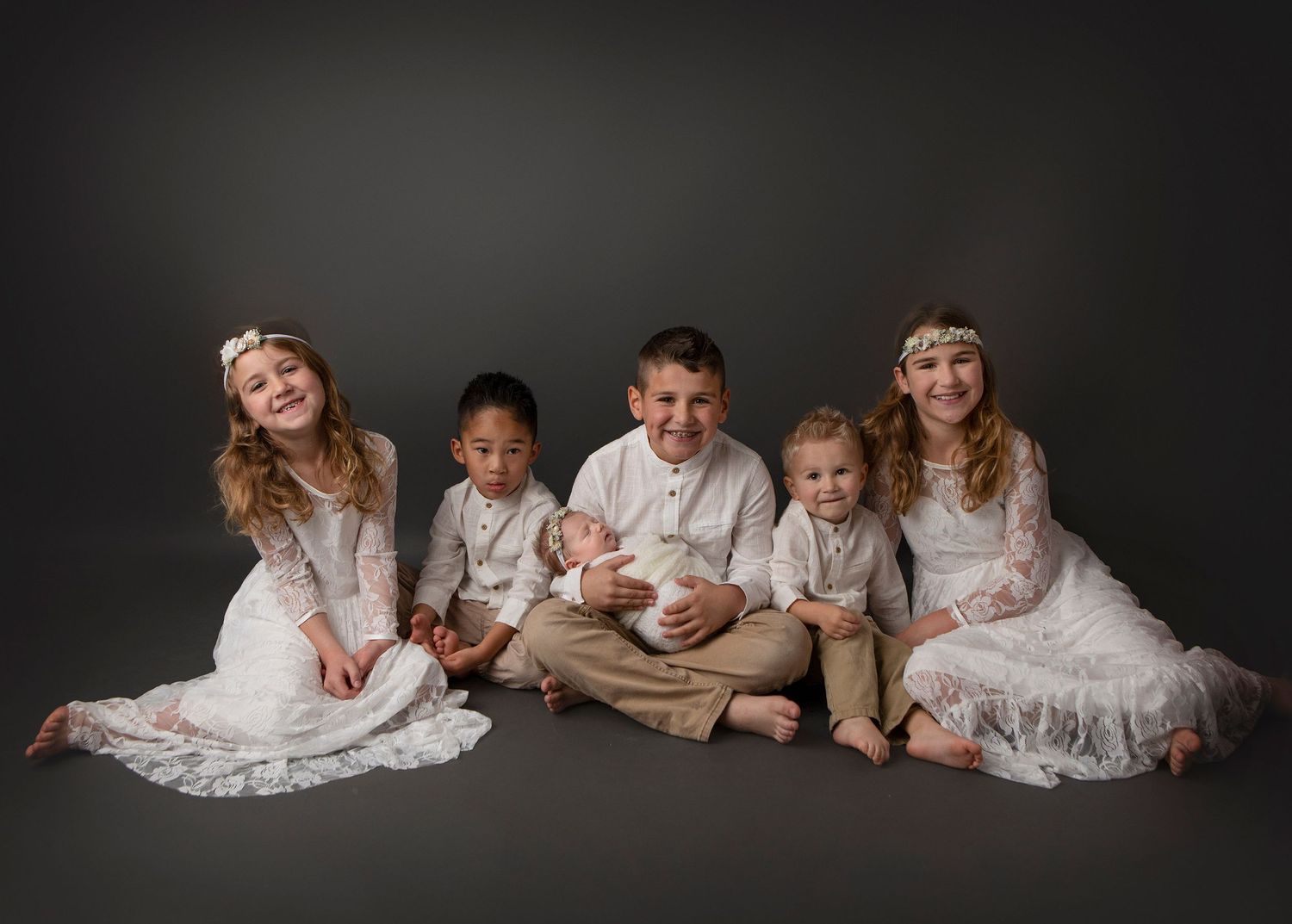 Studio family portrait with siblings in white and neutral tones against dark background.