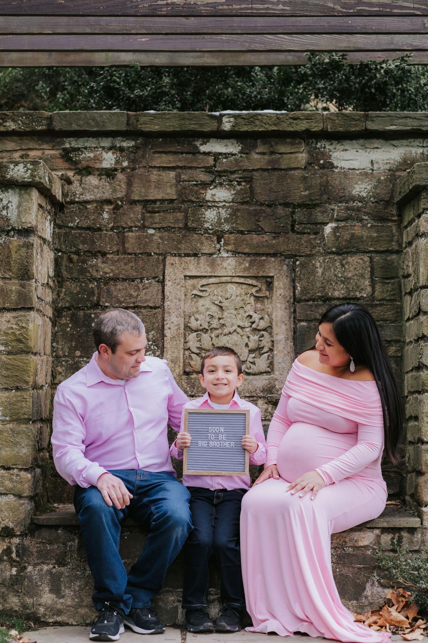 Family maternity photo session in front of stone wall with couple and child holding chalkboard sign.
