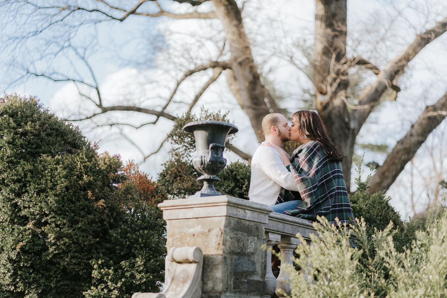 A couple shares an intimate moment on a stone pillar in a garden setting.