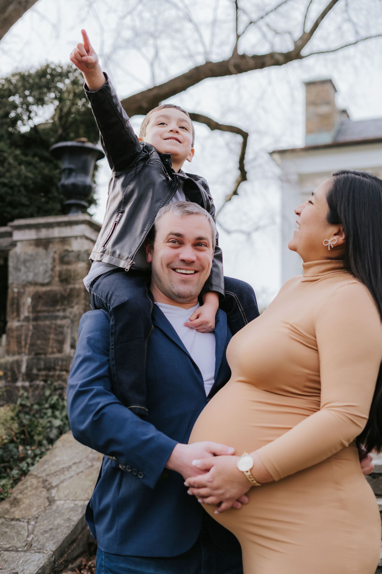 Joyful outdoor family moment with expecting couple and child on shoulders against stone backdrop.