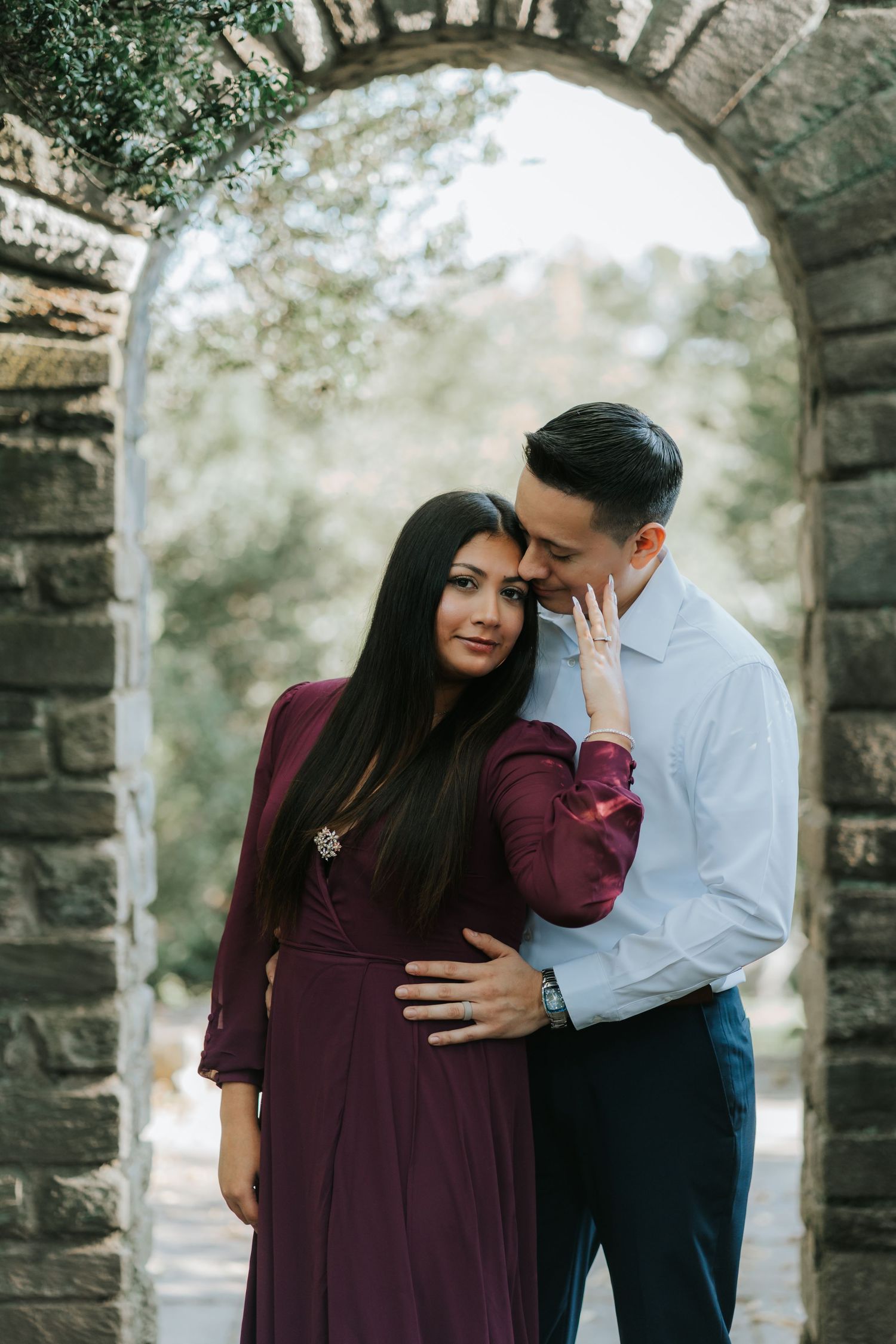 A romantic couple embraces under a stone archway, with her in a burgundy dress and him in a light blue shirt.