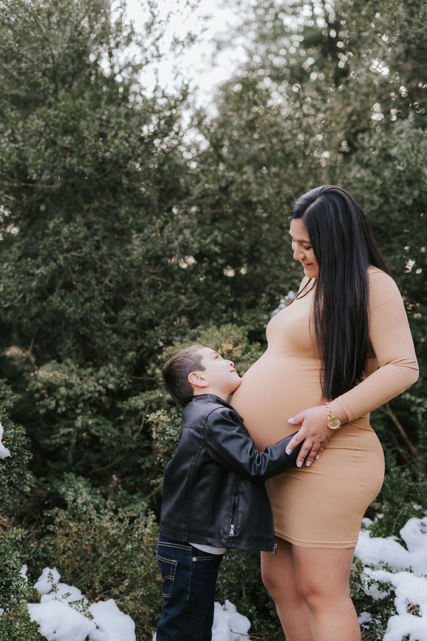 Tender moment between young child and expectant mother in fitted tan dress outdoors.