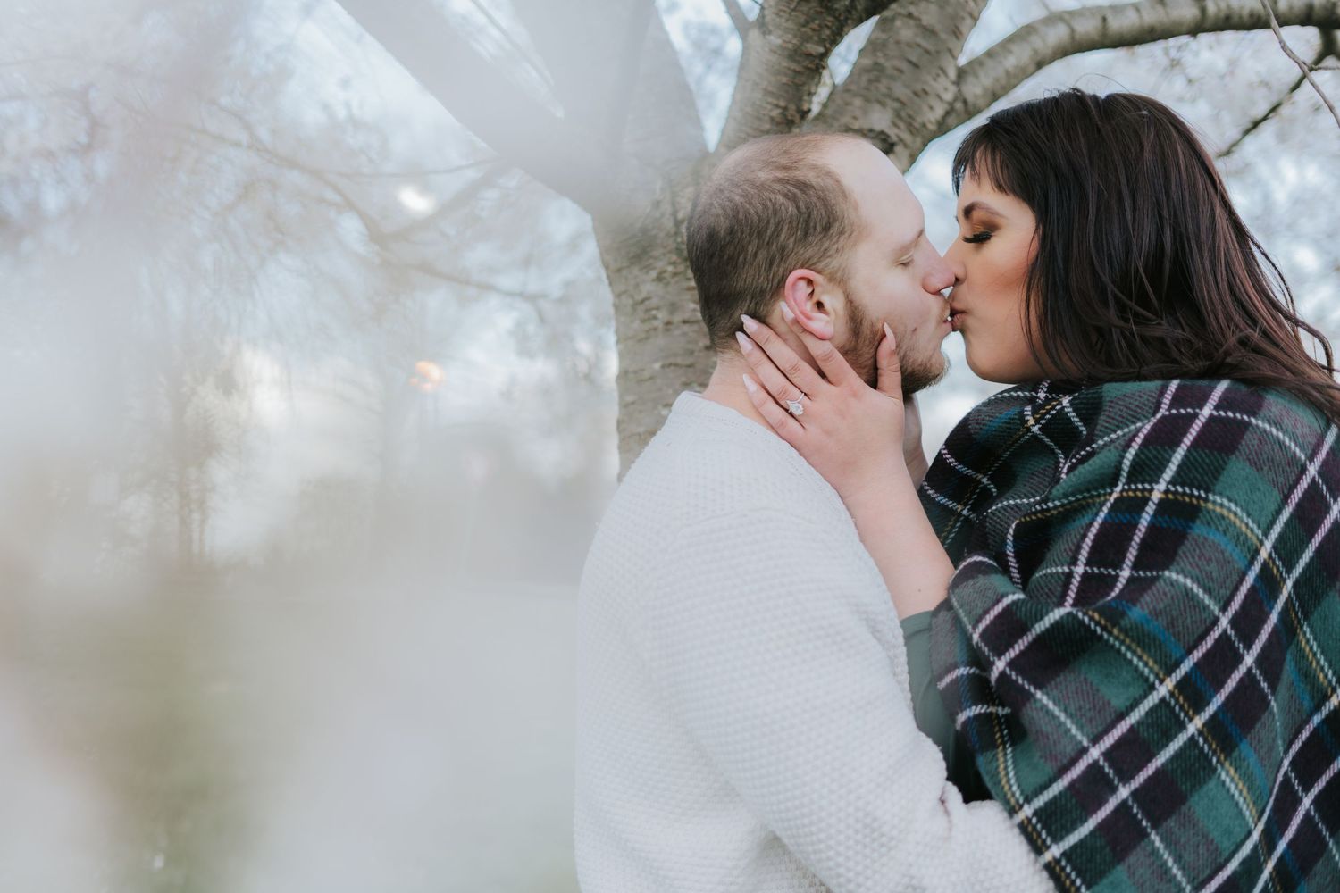 A couple shares a romantic kiss under blooming trees on a misty day.