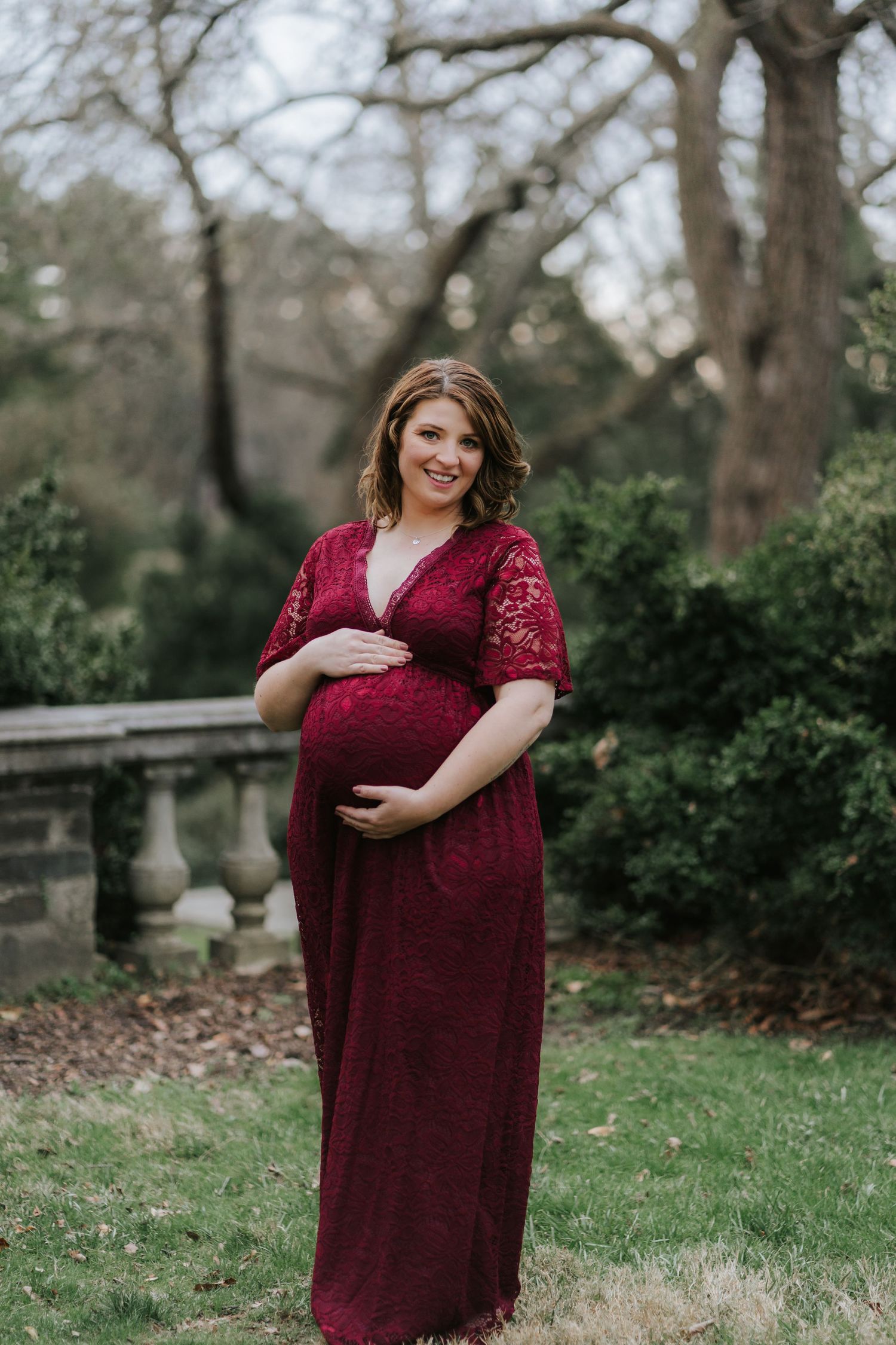 Maternity portrait of a woman in a burgundy lace dress standing in a garden setting.