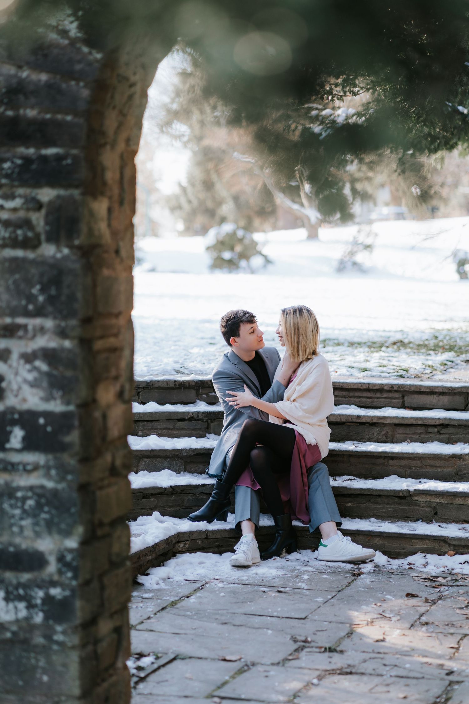 A romantic winter engagement photo of a couple sitting on snowy stone steps framed by an archway.