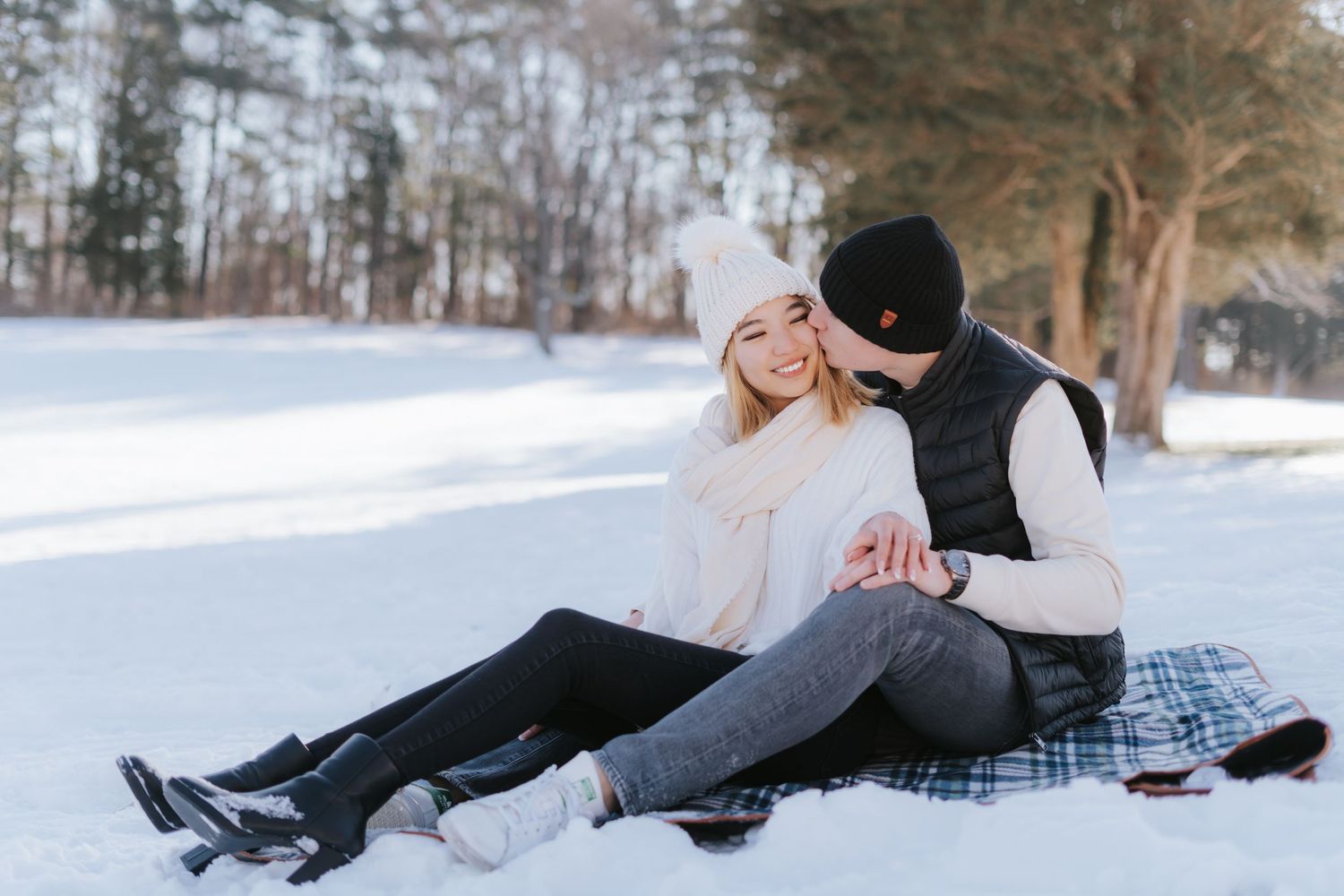 A couple sharing an intimate moment while sitting on a plaid blanket in the snow.