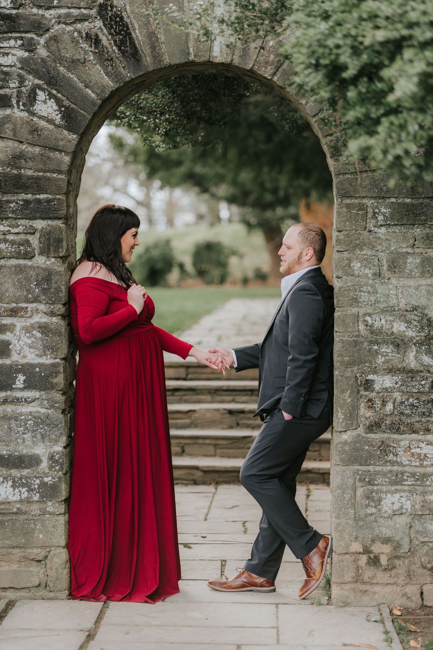 A couple in red dress and grey suit poses playfully under a historic stone archway.
