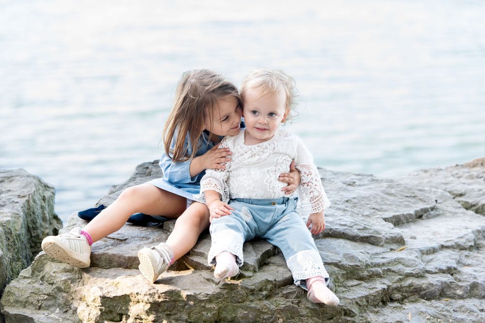 Two children sitting closely together on rocks near waterfront.